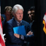 Clinton CRASHES Cowboys Draft—Jones Nearly Topples… Bill Clinton standing between American flags with a folder.