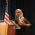 Woman speaking at podium with American flag background.