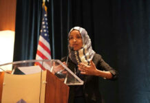 Omar Stays Put — D.C. DRAMA Ahead… Woman speaking at podium with American flag background.