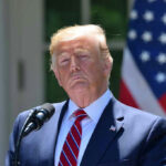 Trump’s Wreath-Laying CEREMONY: Apolitical or Not? Man speaking at podium with American flag background.