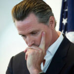 Newsom’s Controversial Boycott Draws STINGING Criticism… Man in suit thinking with hand on face near a U.S. flag