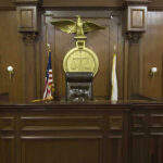 Mail-Order Death Dealer Walks on Murder Judges bench in an empty courtroom, scales emblem above.