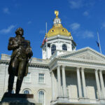 Race For Lindsey Graham’s Seat Turns Volatile New Hampshire State House with statue in foreground.