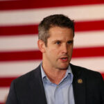 Flag Jacket Ban Ignites Outrage Man speaking in front of American flag backdrop.