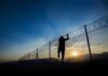 Silhouette of person climbing a wire fence at sunset.