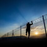 Silhouette of person climbing a wire fence at sunset.